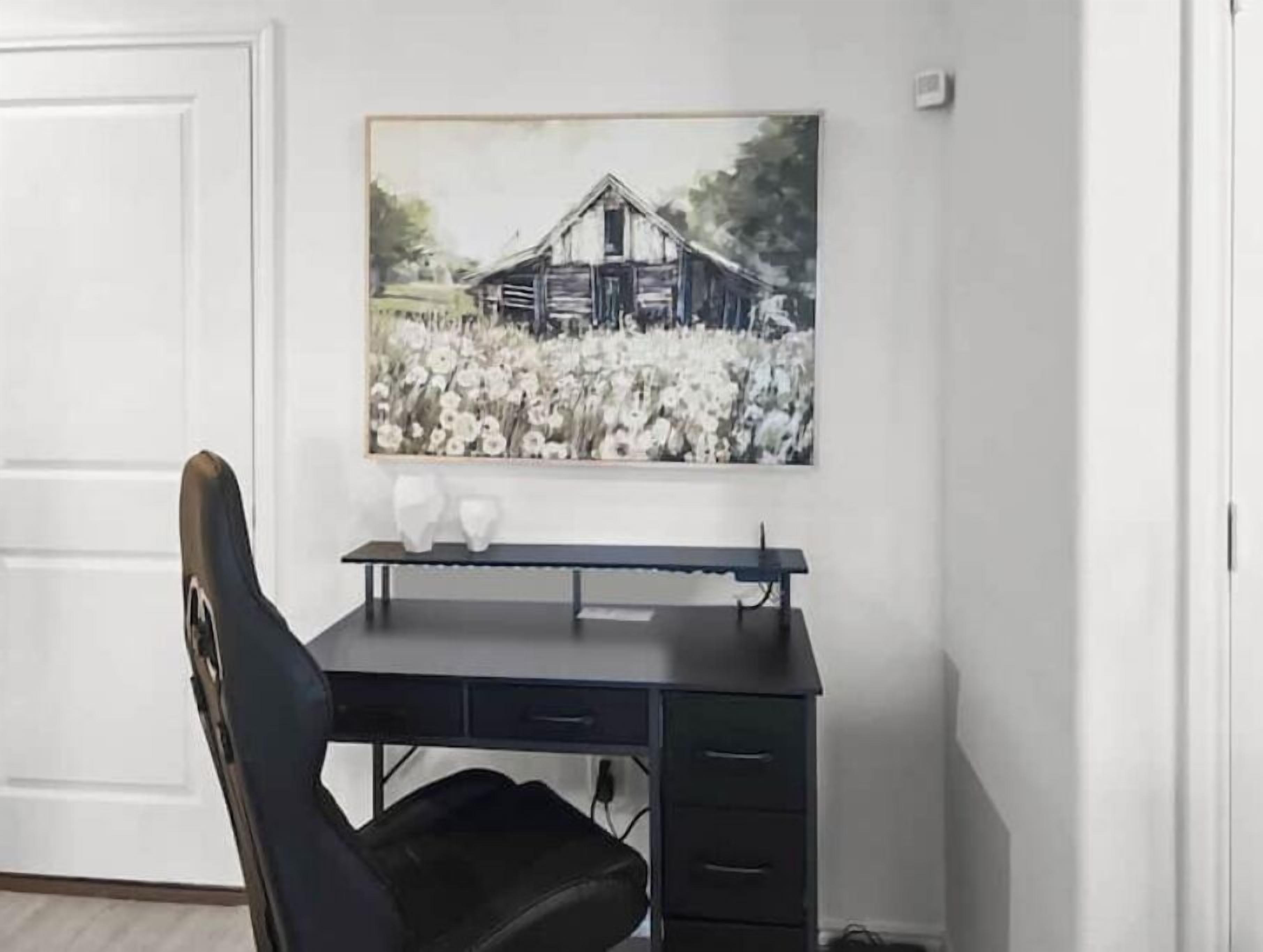 A room with a desk, a chair, and a painting of a barn and flowers on the wall.