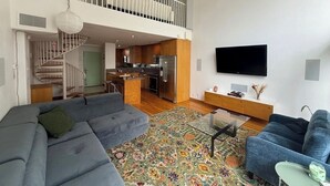 Ein großzügiger Wohnraum mit einer großen grauen Couch, einer blauen Couch und einem farbenfrohen Teppich. Eine Treppe führt zu einer oberen Ebene mit einem Balkon.