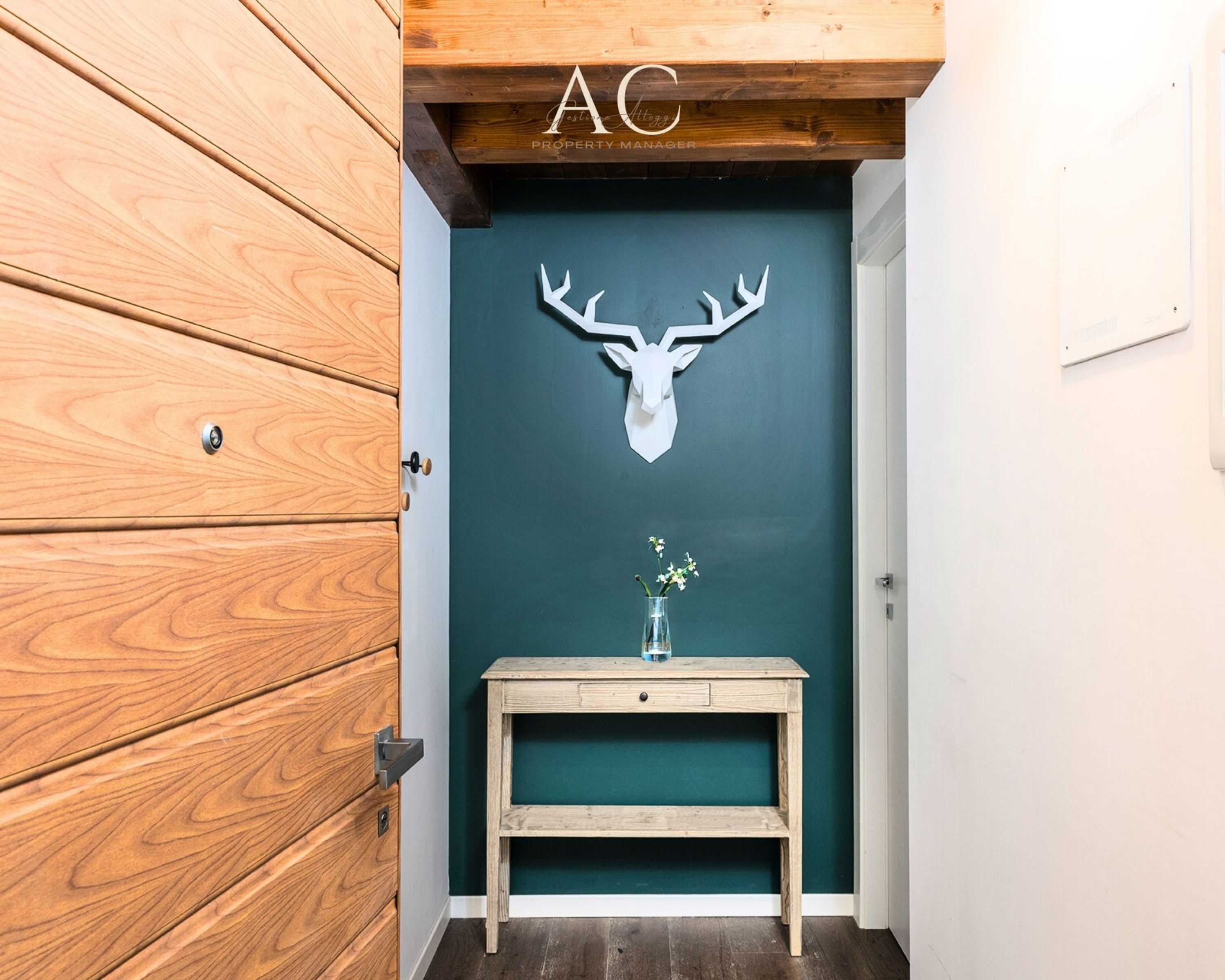 A wooden door with a mounted deer head, a small wooden table with a vase, and a teal wall.