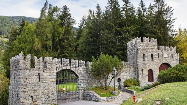 Entrada de un castillo de piedra con un camino adoquinado que lo lleva al interior, rodeado de árboles y una colina cubierta de césped.