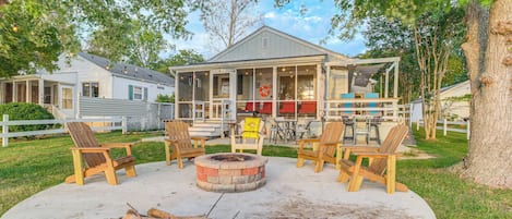 A covered outdoor seating area with wooden chairs and a central fire pit, surrounded by a grassy lawn and trees.