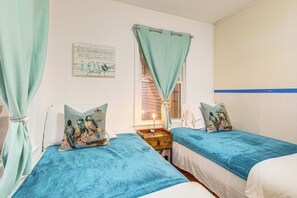 A bedroom with two single beds, each with a blue bedspread and matching pillows. There is a small wooden nightstand between the beds, and a window with curtains. - Potomac River Getaway: Walk to Marina! (King George)