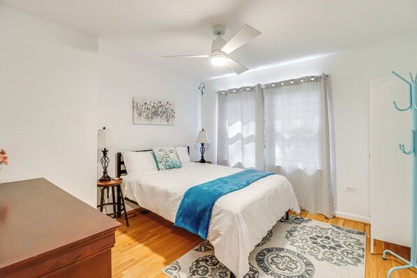 Un dormitorio con cama, ventilador de techo, mesita de noche, lámpara, ventana con cortinas y perchero.