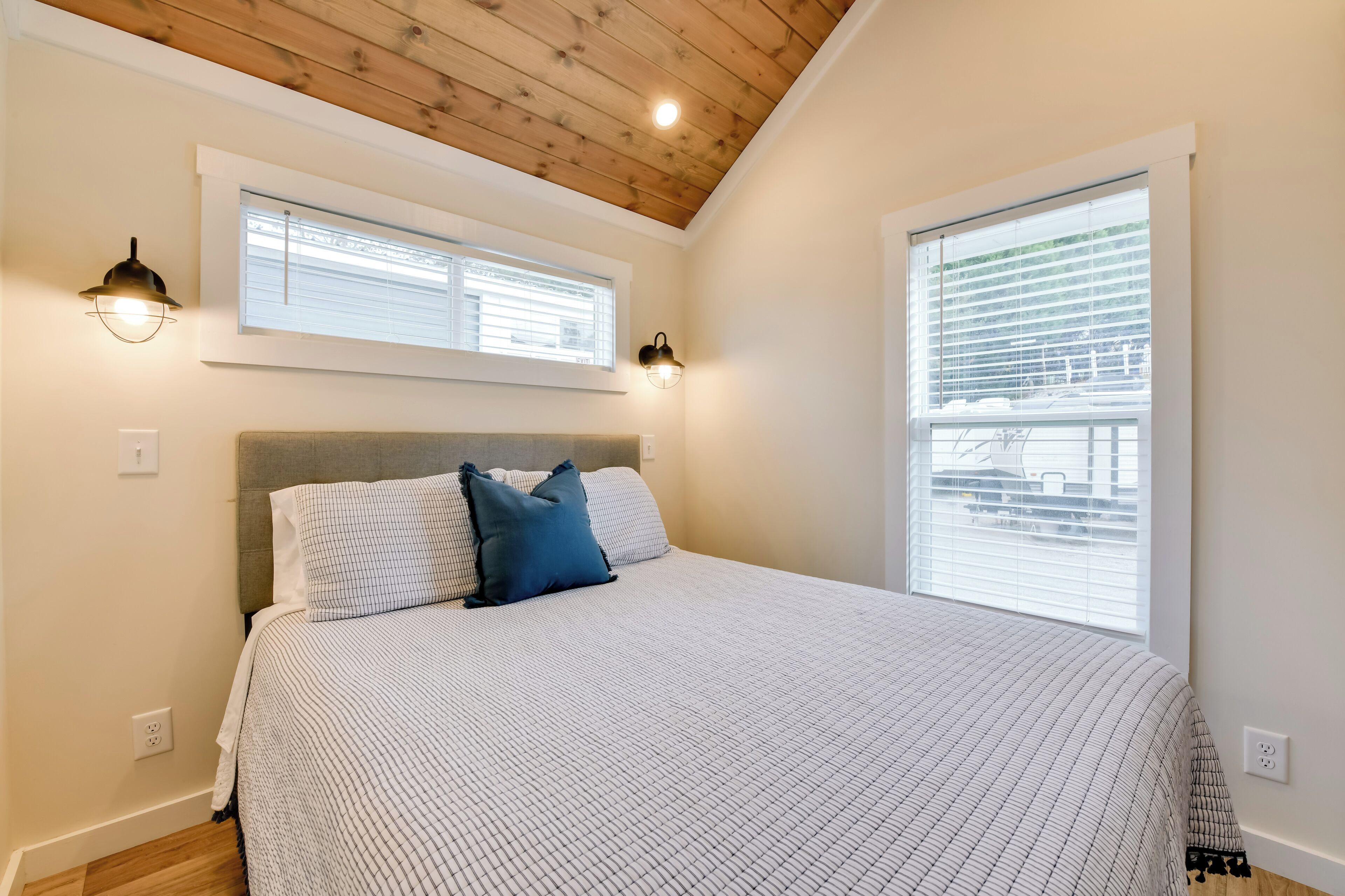Een slaapkamer met een houten plafond, een groot bed met een beddengoed met een patroon, twee wandlampen en een raam met jaloezieën.