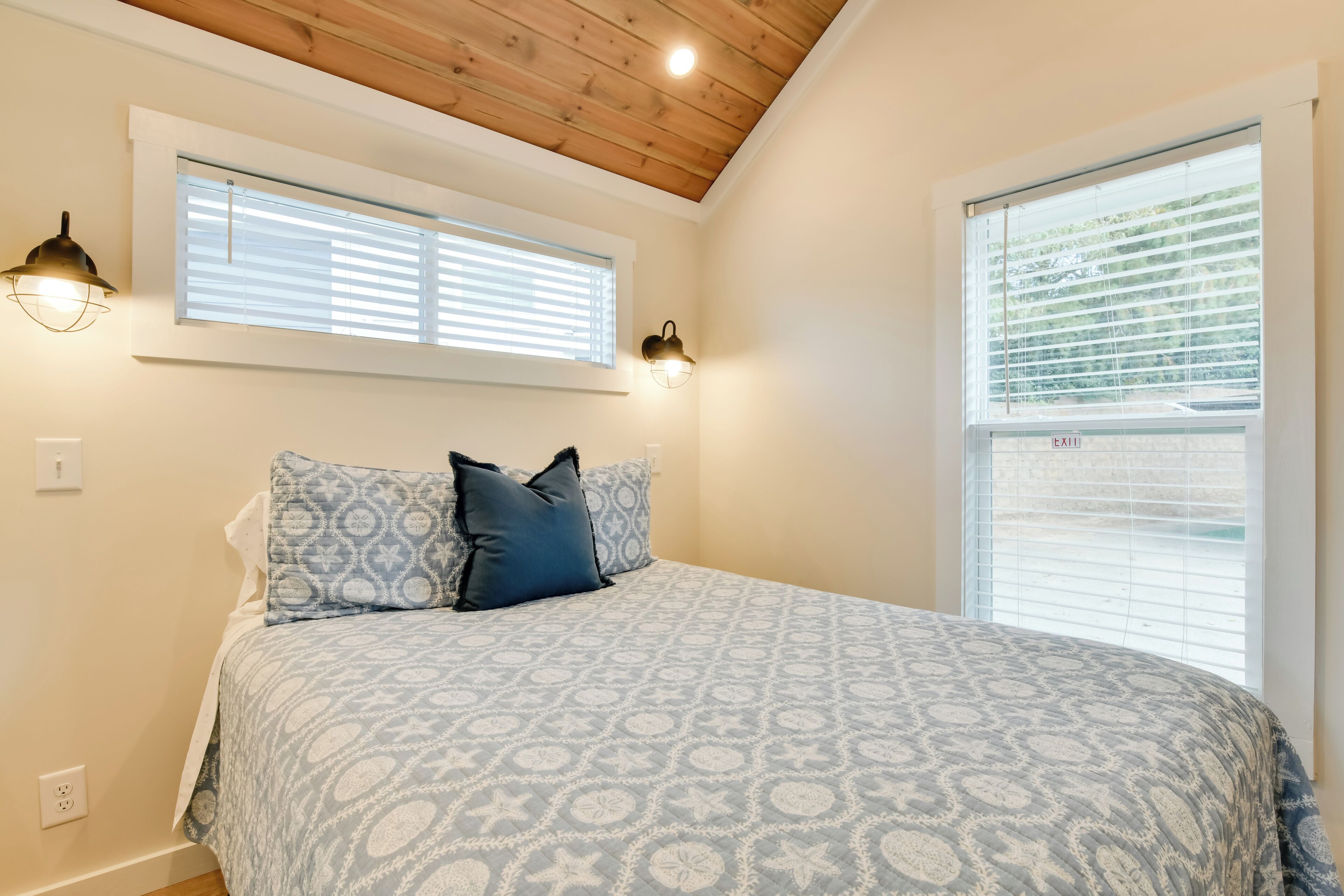 A bedroom with a patterned bedspread, two wall-mounted lamps, a window with blinds, and a ceiling with wooden beams.
