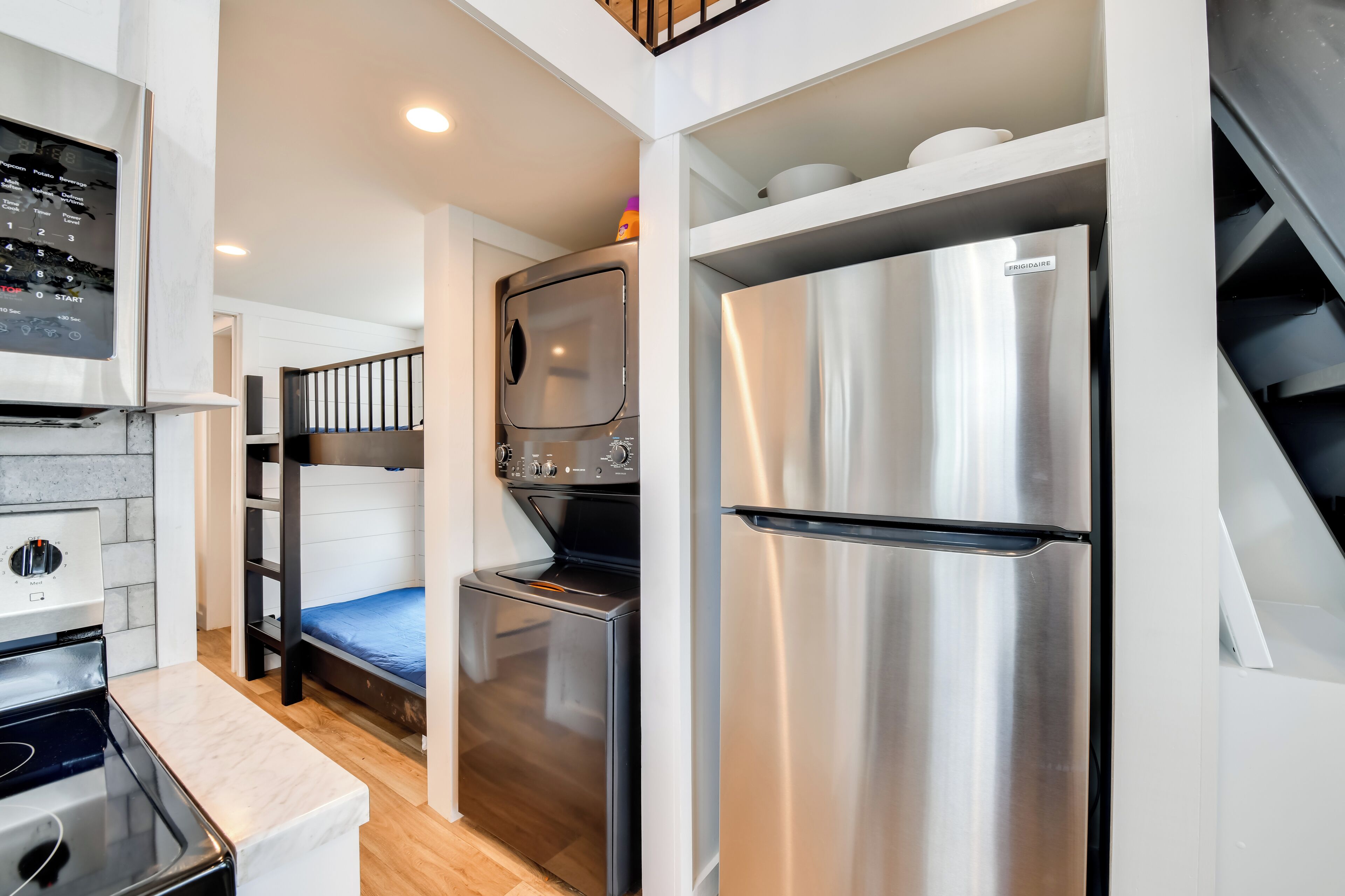 A compact kitchen with a built-in oven, a stainless steel refrigerator, and a microwave. There is a bunk bed with a blue mattress in the background.