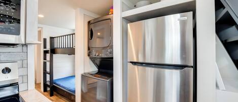 A compact kitchen with a built-in oven, a stainless steel refrigerator, and a microwave. There is a bunk bed with a blue mattress in the background.
