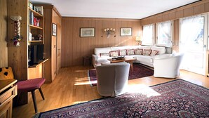 Amplia sala de estar con un sofá grande, una mesa de centro y una alfombra estampada. Se observa una estantería con libros y objetos decorativos, además de un mueble de madera.