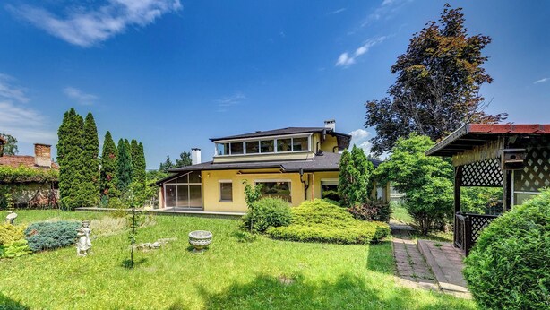 Une maison à deux étages avec un jardin à l’avant, un ciel bleu dégagé et un arbre sur la droite.