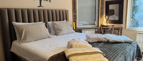 A neatly made bed with pillows and a blanket, a wall-mounted air conditioner, a window with a view, and a small table with a chair.