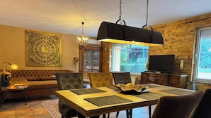 Ein Essbereich mit einem Holztisch, vier Stühlen und einer Kronleuchter. Im Hintergrund befindet sich eine Couch und ein Fernseher.