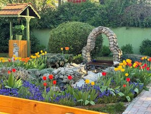 Ein Garten mit einem steinernen Torbogen, einem hölzernen Gartenpavillon und verschiedenen Blumen, darunter Tulpen und violette Blüten.