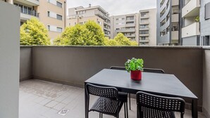 Un balcon extérieur avec une table et des chaises, un pot de fleurs et une vue sur les immeubles d’habitation environnants.