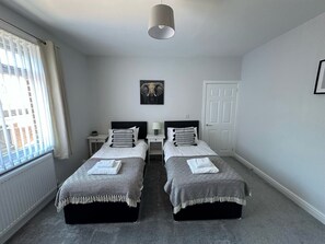 A bedroom with two beds, a radiator, a window with blinds, a door, and a ceiling light. - Storey Apartment - Downstairs 2bed Flat (Newbiggin-by-the-Sea)