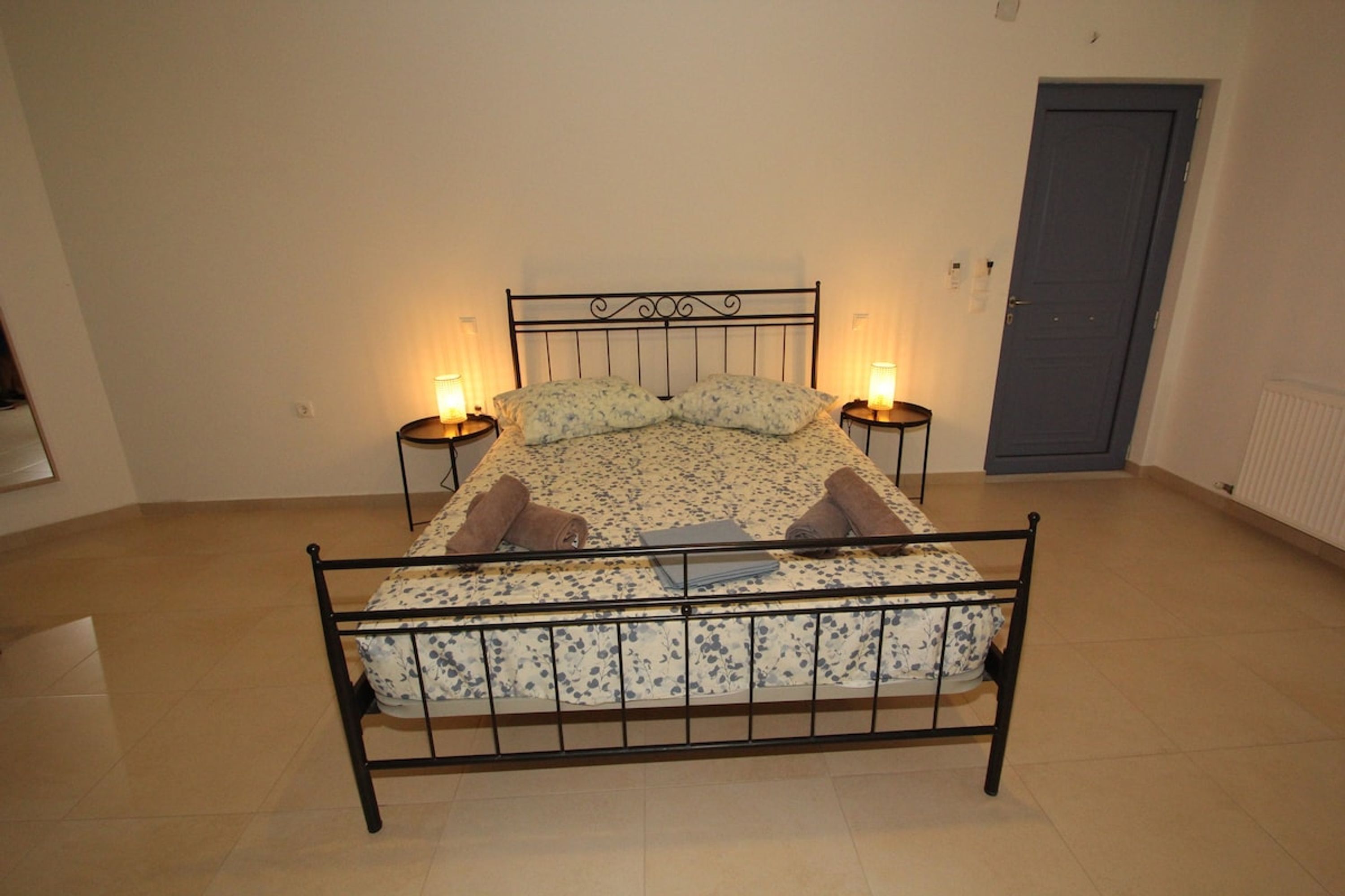 A single bed with a metal frame, two bedside tables, and a door in a room with plain walls and tiled flooring.