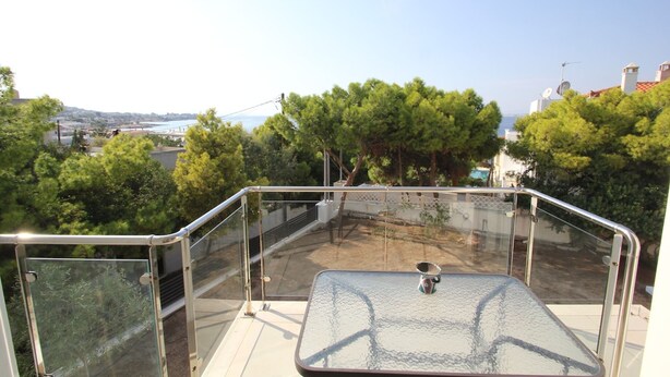 A balcony with a glass table and metal railing, overlooking a residential area with trees and a distant bridge. - Schinos by Frangki (Aegina)