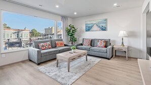 A modern living room with two grey sofas, a wooden coffee table, a large window with a view of buildings, and a painting of a pier. - 3-bedroom house on 2F in peaceful Daly City with AC, WiFi. (Daly City)