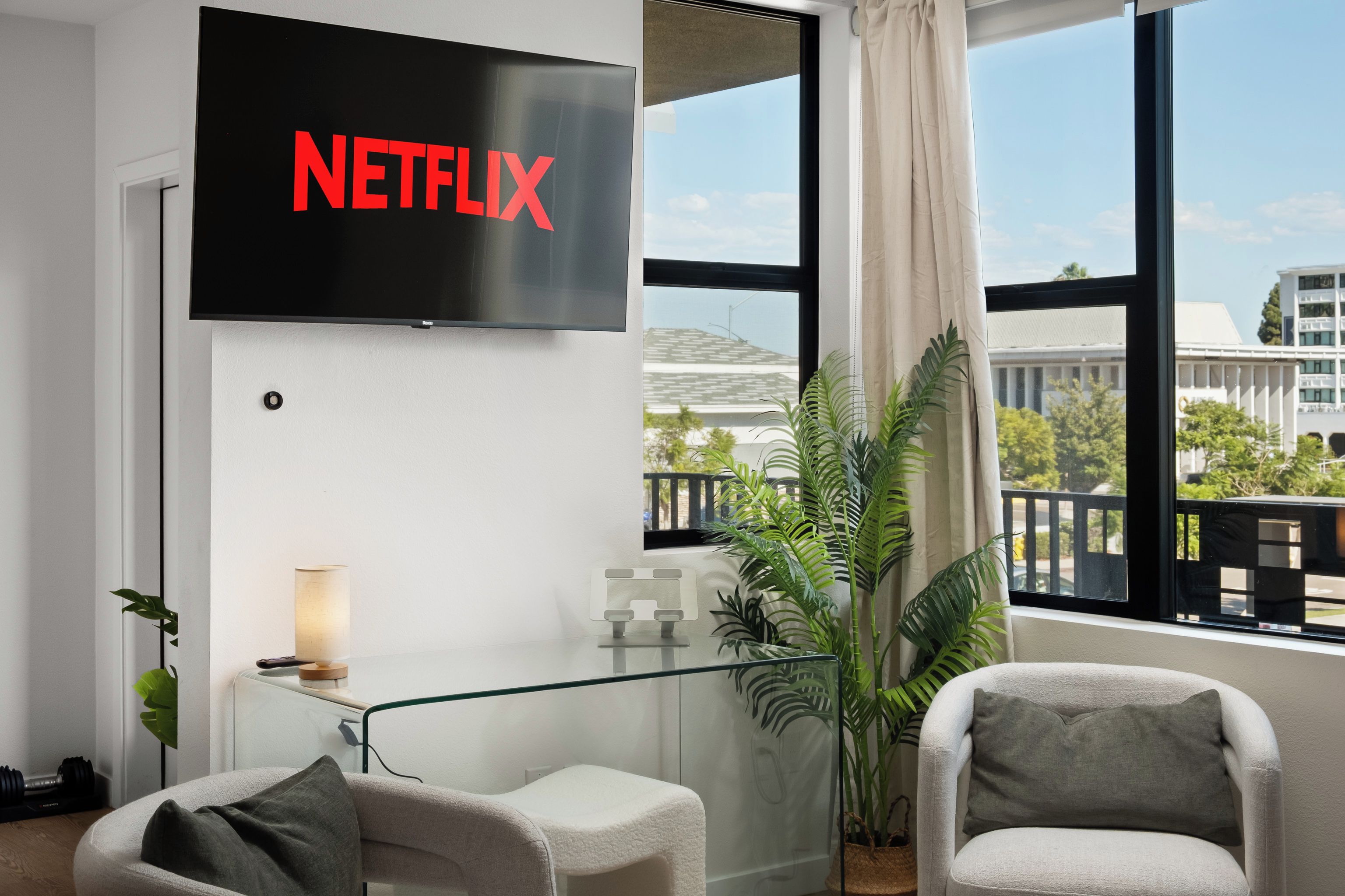 A modern living room with a large flat-screen TV mounted on the wall displaying the Netflix logo, two armchairs, a small table with a lamp, and a large window with a view of a building and greenery outside.