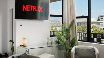 A modern living room with a large flat-screen TV mounted on the wall displaying the Netflix logo, two armchairs, a small table with a lamp, and a large window with a view of a building and greenery outside.