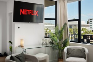 Ein modernes Wohnzimmer mit einem großen Flachbildfernseher an der Wand, auf dem das Netflix-Logo zu sehen ist, zwei Sessel, ein kleiner Tisch mit einer Lampe und ein großes Fenster mit Blick auf ein Gebäude und Grünflächen.