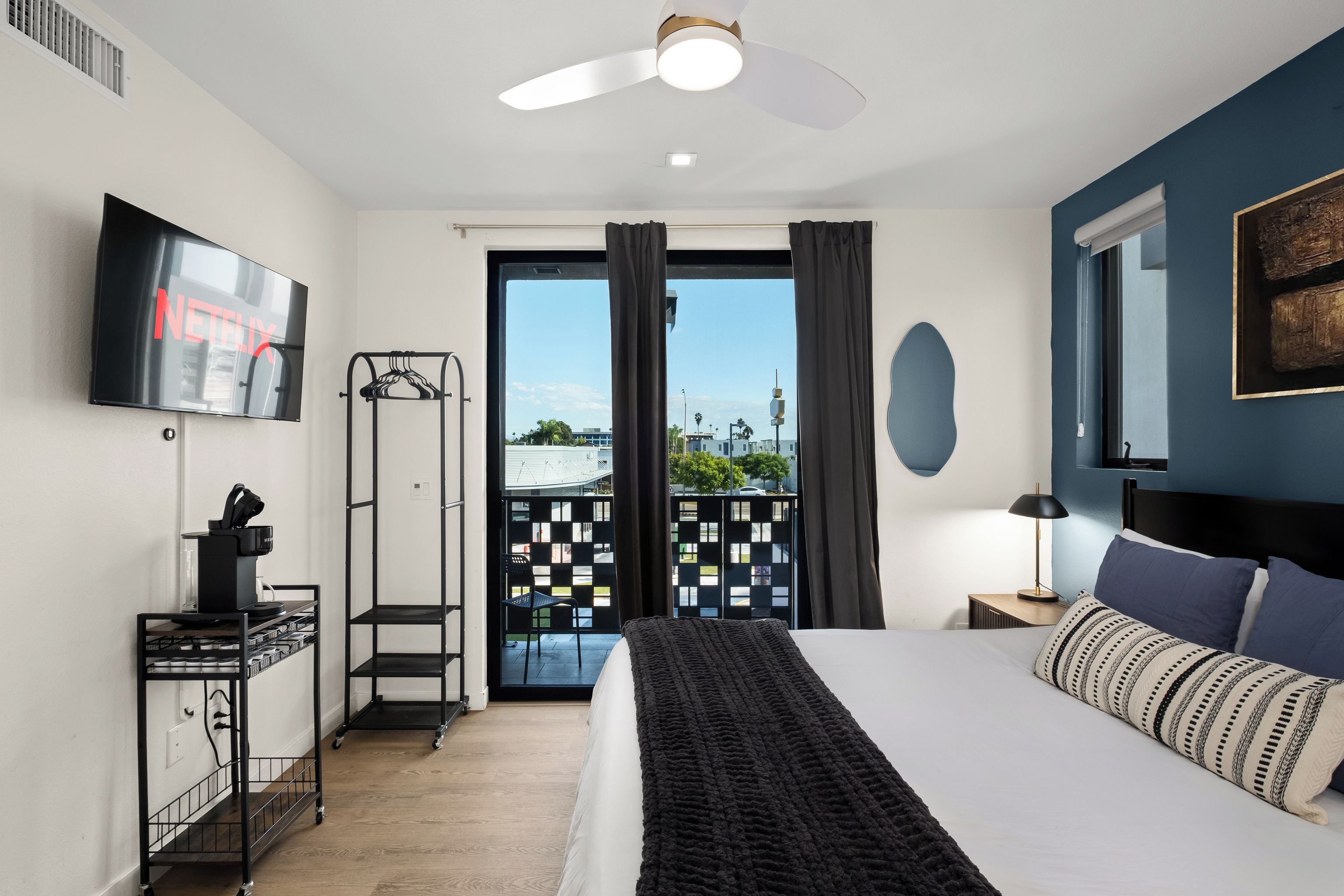 Et hotelværelse med en seng, et tv ophængt på væggen, et skrivebord med en computer og hovedtelefoner, en balkon med udsigt og en ventilator i loftet.