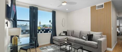 A modern living room with a sofa, coffee table, and TV, a balcony with curtains, and a city view.