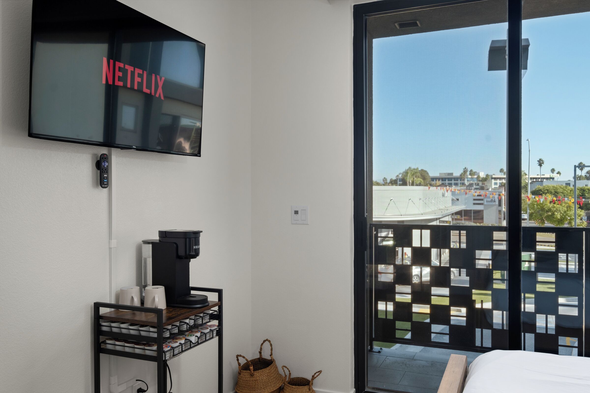 Une chambre d’hôtel équipée d’un téléviseur à écran plat fixé au mur, sur lequel apparaît le logo de Netflix, d’une machine à café posée sur une étagère et offrant une vue sur l’extérieur grâce à une porte-fenêtre coulissante.