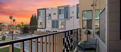 A modern building with a balcony, outdoor seating, and a view of the street at sunset.