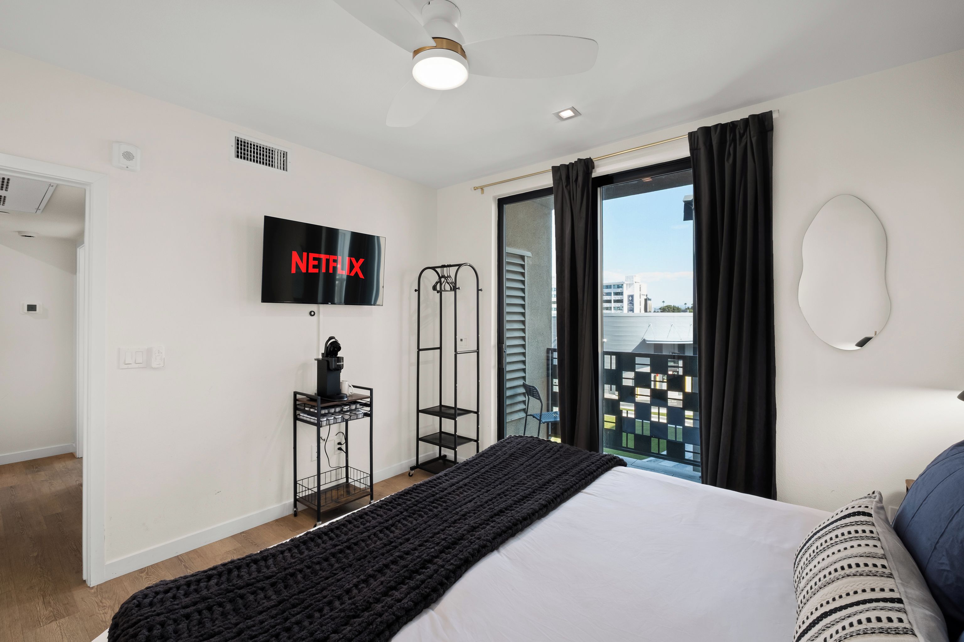 A modern hotel room with a bed, a TV mounted on the wall, a ceiling fan, and a view of the city through the window.