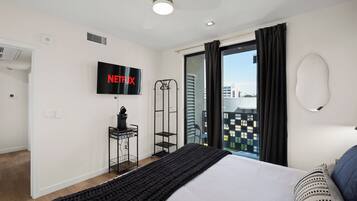 A modern hotel room with a bed, a TV mounted on the wall, a ceiling fan, and a view of the city through the window.