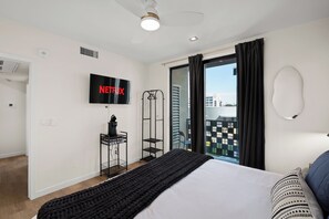Una habitación de hotel moderna con una cama, un televisor montado en la pared, un ventilador de techo y vistas a la ciudad a través del ventanal.