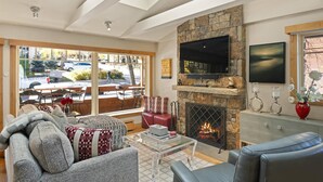 A modern living room with a stone fireplace, a flat-screen TV, and a large window overlooking a street. - Fasching Haus 03 (Aspen)