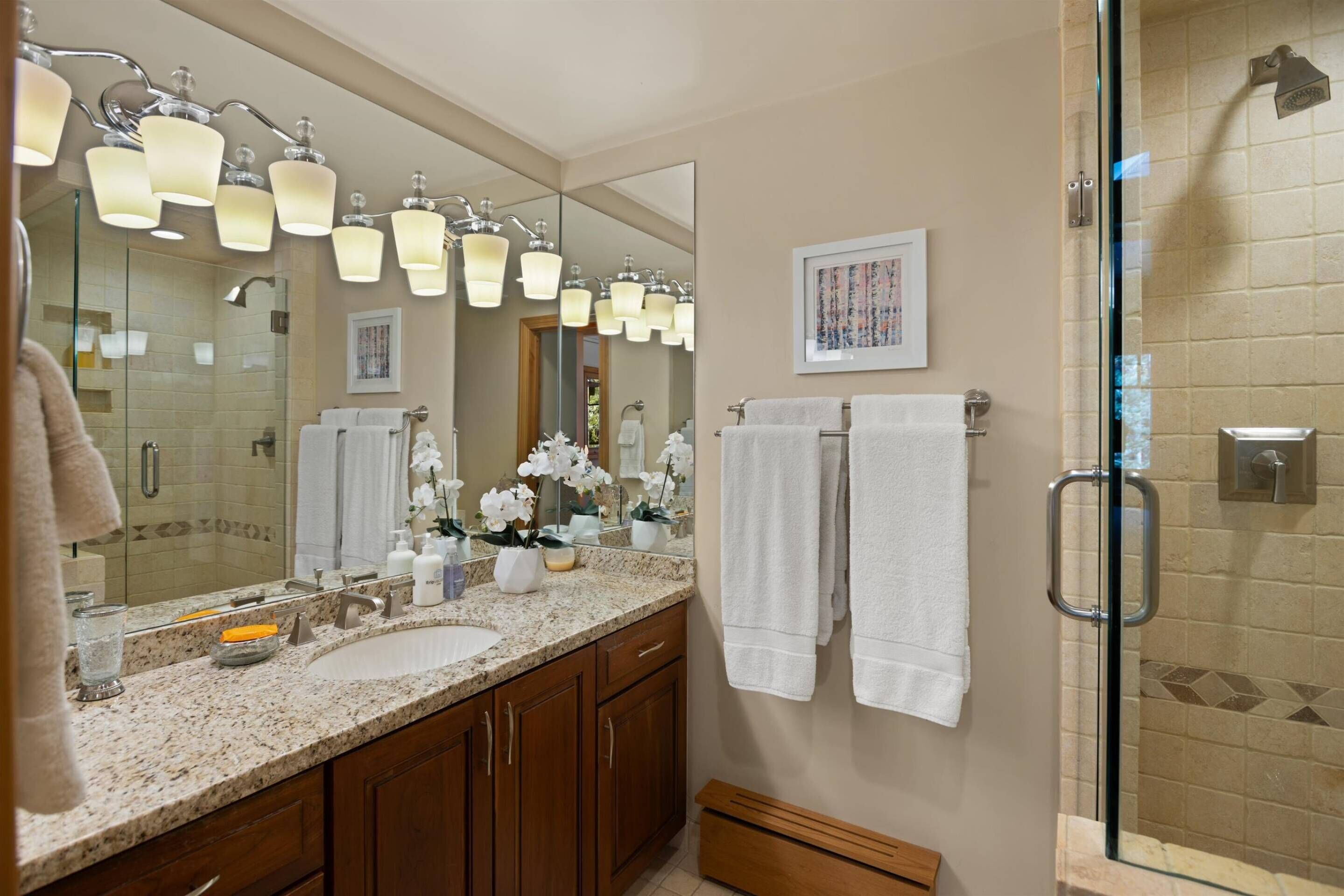 A bathroom with a large mirror, multiple light fixtures, a sink, and a shower area.