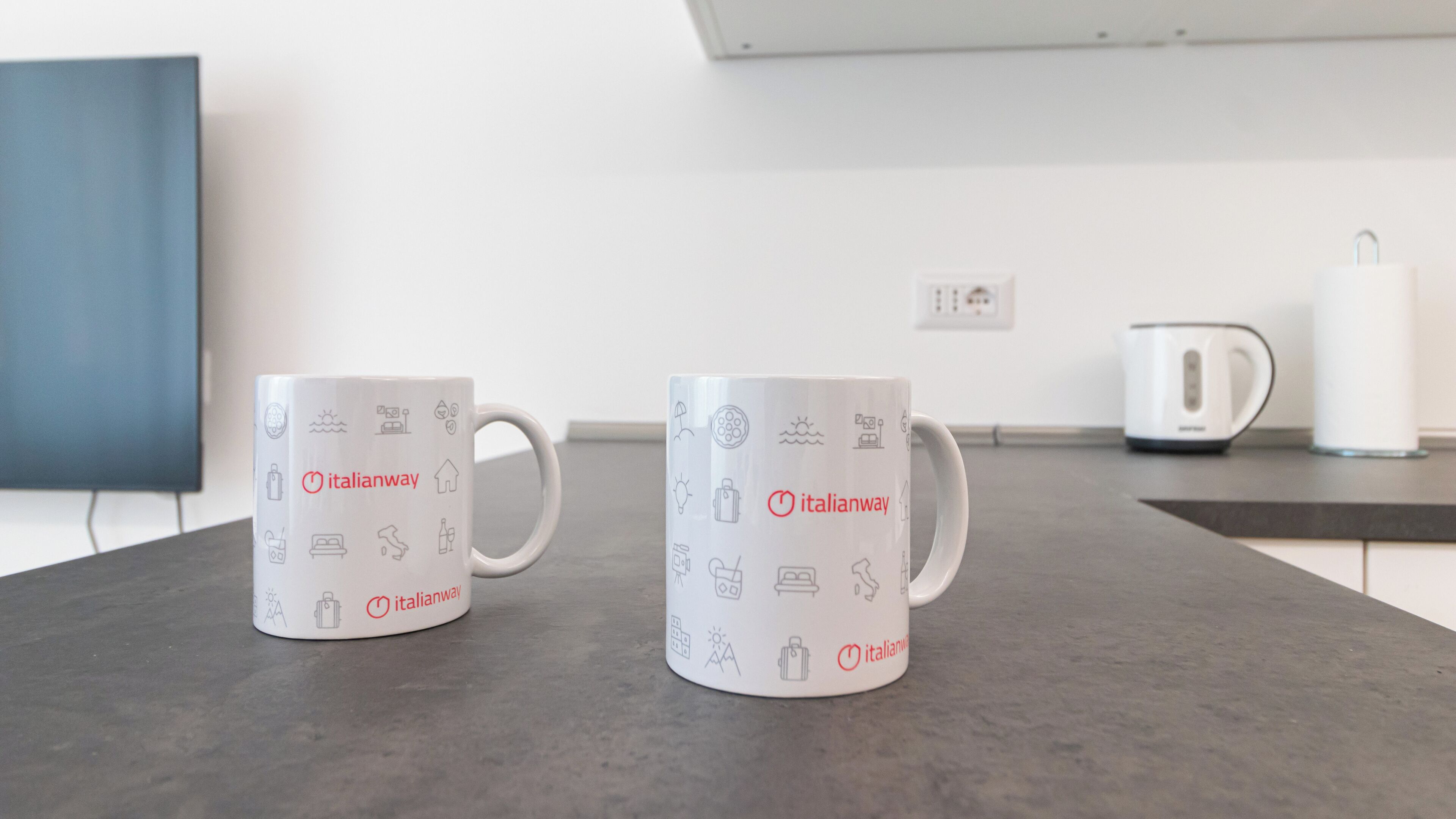 Deux tasses blanches ornées du logo Italianway et de motifs liés au voyage, posées sur un plan de travail de cuisine.