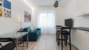 A modern, well-lit room with a glass dining table, a sofa with colorful cushions, a TV on a stand, and a kitchen area with cabinets and a countertop. - Italianway Comfort - Serra 16 (Milano)
