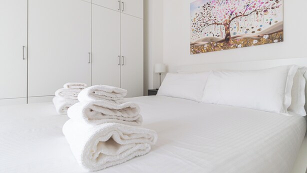 A neatly made bed with white linens and a stack of folded towels on the side. - Italianway Comfort - Serra 16 (Milano)