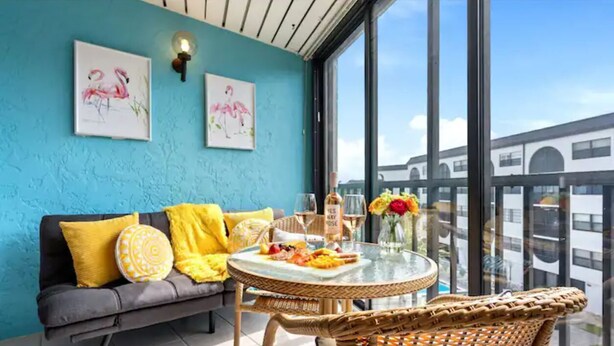 Un balcon avec un fauteuil en osier, une table en verre ornée d’un vase de fleurs et un canapé avec des coussins jaunes.