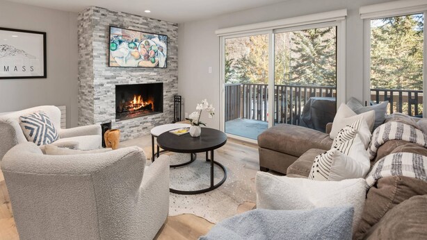 A cozy living room with a fireplace, comfortable seating, and a balcony with a view of trees.