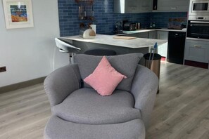 Una sala de estar moderna con un sillón gris, una almohada rosa y una cocina al fondo.