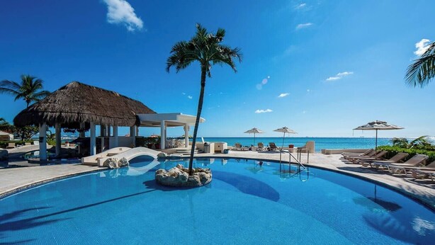 Een resort zwembad met ligbedden, een palmboom en een overkapping met rieten dak.