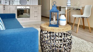 Uma sala de estar moderna com um sofá azul, uma mesa de centro redonda de fibra natural e uma área de jantar com uma cadeira branca e um vaso.