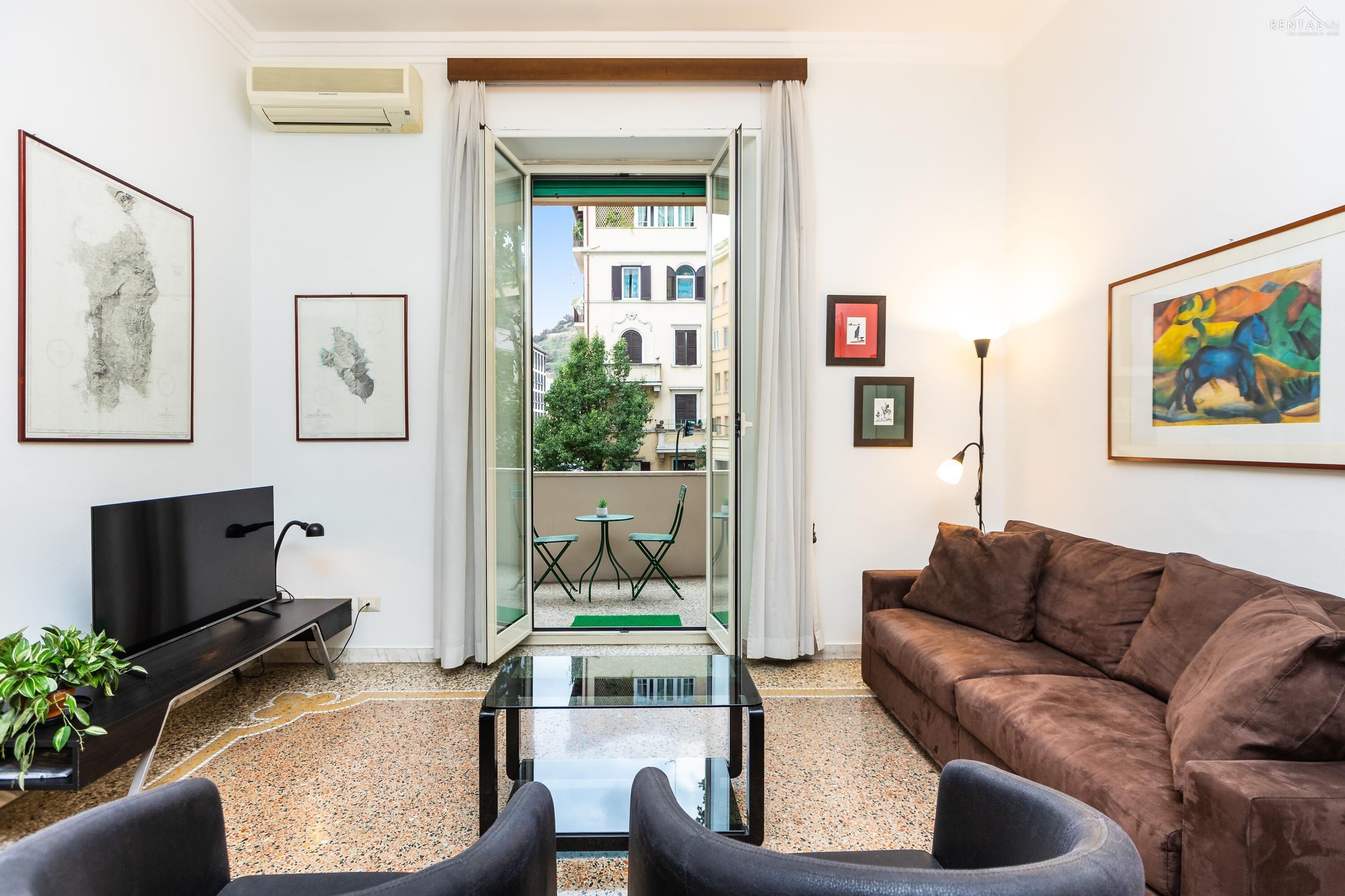 Ein modernes Wohnzimmer mit einer Couch, einem Glastisch und einem Fernseher. An den Wänden hängen gerahmte Kunstwerke und vom Balkon aus hat man einen Blick auf die Häuser.