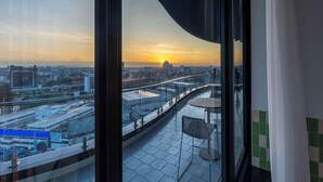 Un balcón con vistas a la ciudad al atardecer, barandilla de cristal y una mesa con sillas.