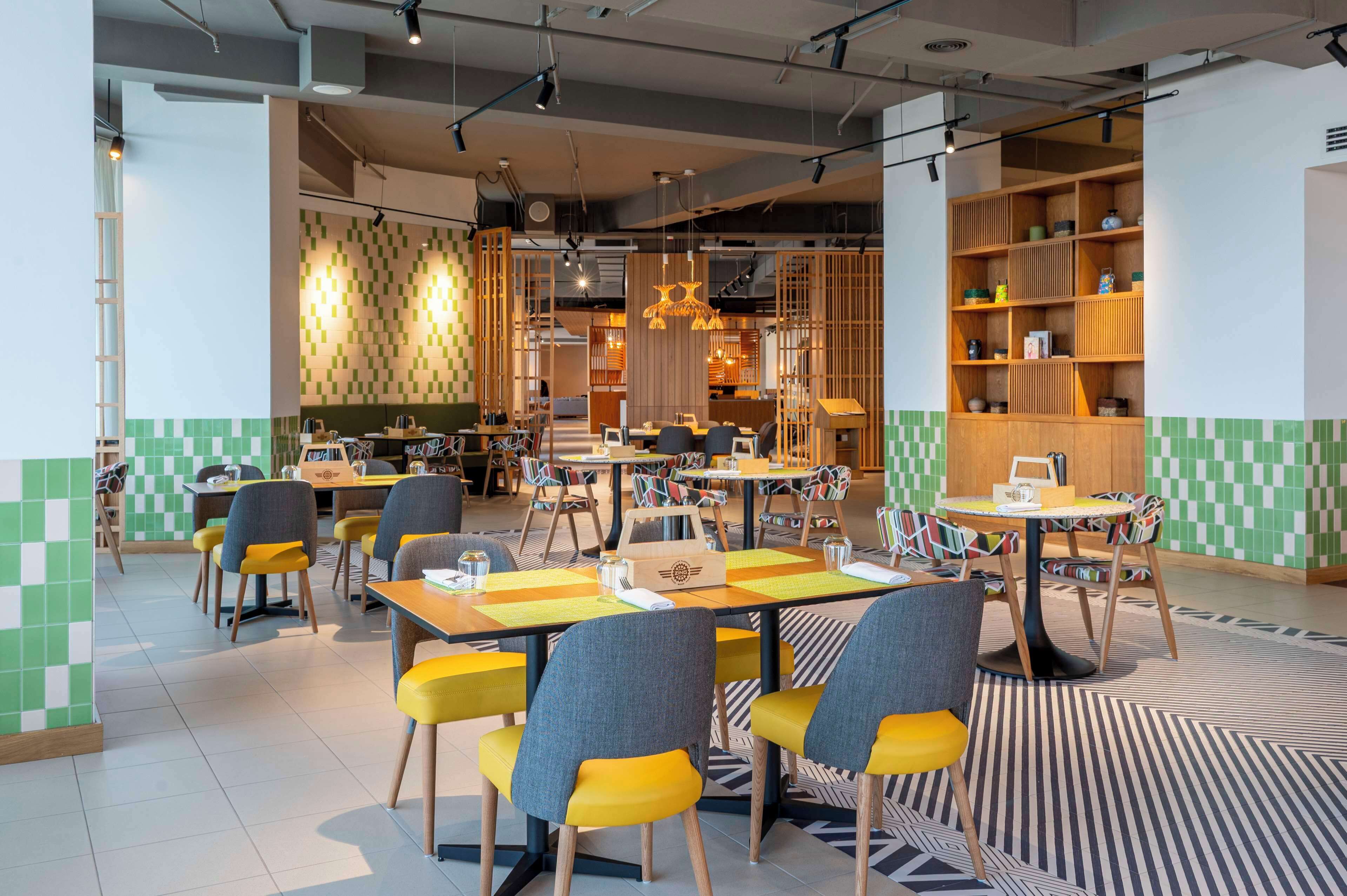En moderne restaurant med flere spiseområder, herunder et centralt åbent område med borde og stole, samt et afskilt spiseområde med en træbogreol.