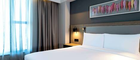 Habitación de hotel con una cama grande, una mesita de noche con lámpara, una obra de arte en la pared y cortinas translúcidas.