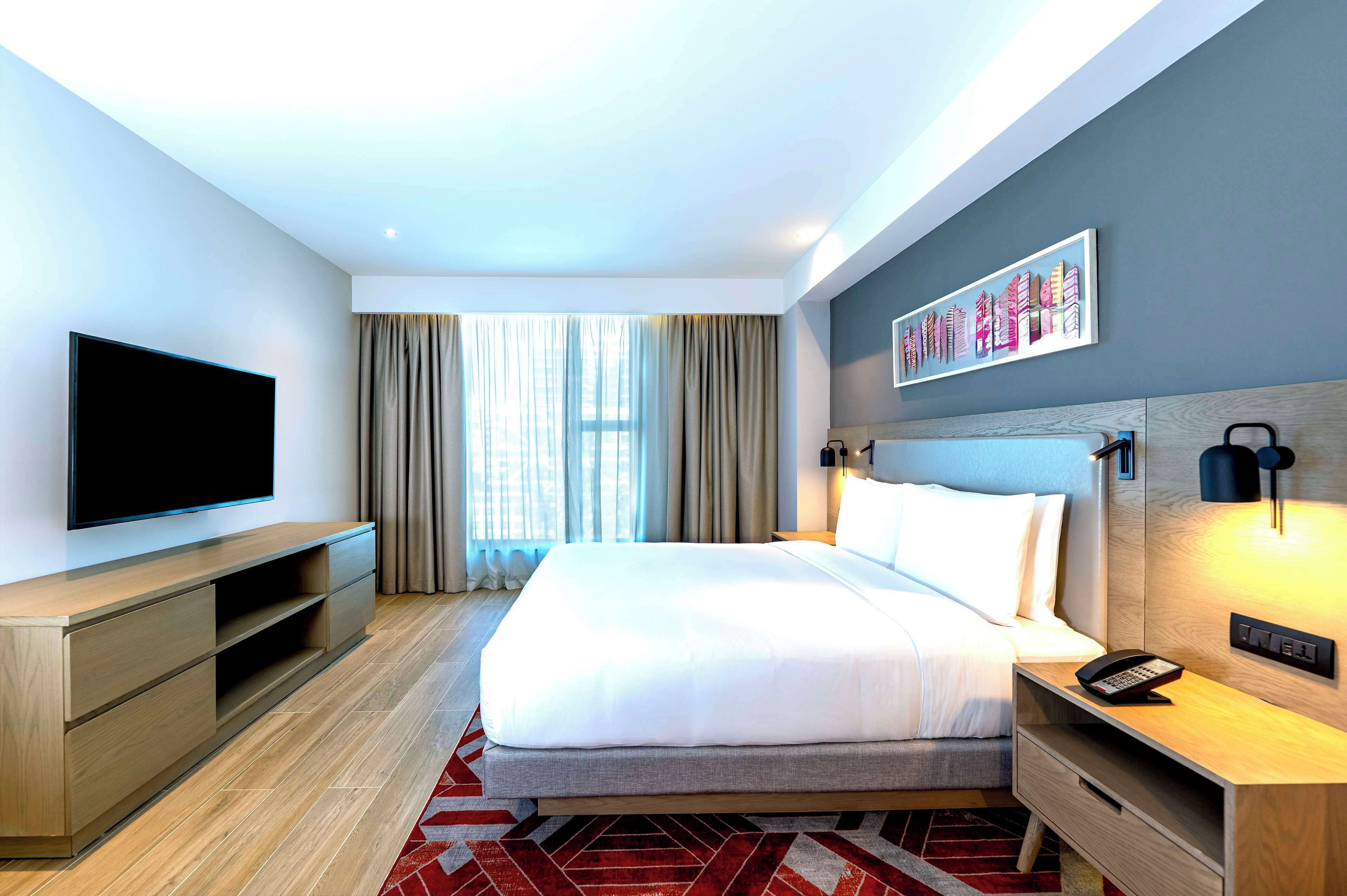 A modern hotel room with a large bed, a flat-screen TV, a wooden nightstand, and a wall-mounted light.