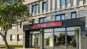 Une entrée d'hôtel moderne avec une façade en verre, une enseigne rouge au-dessus des portes et un arbre sur le côté gauche.