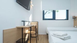 Une chambre d’hôtel moderne équipée d’un lit, d’un bureau avec un ordinateur portable, d’une chaise, d’un téléviseur fixé au mur et d’une fenêtre donnant sur des arbres.