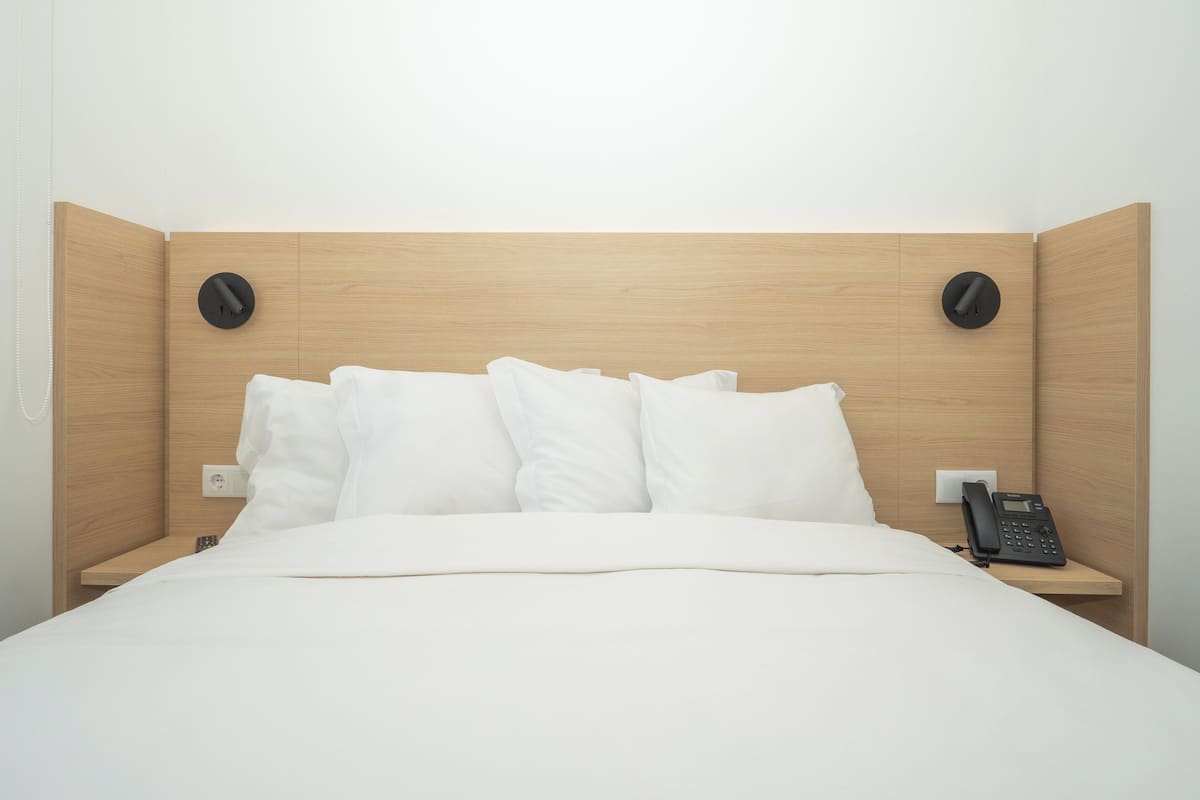 Moderni hotellihuone, jossa on puinen sängynpääty, valkoiset vuodevaatteet ja puhelin yöpöydällä.