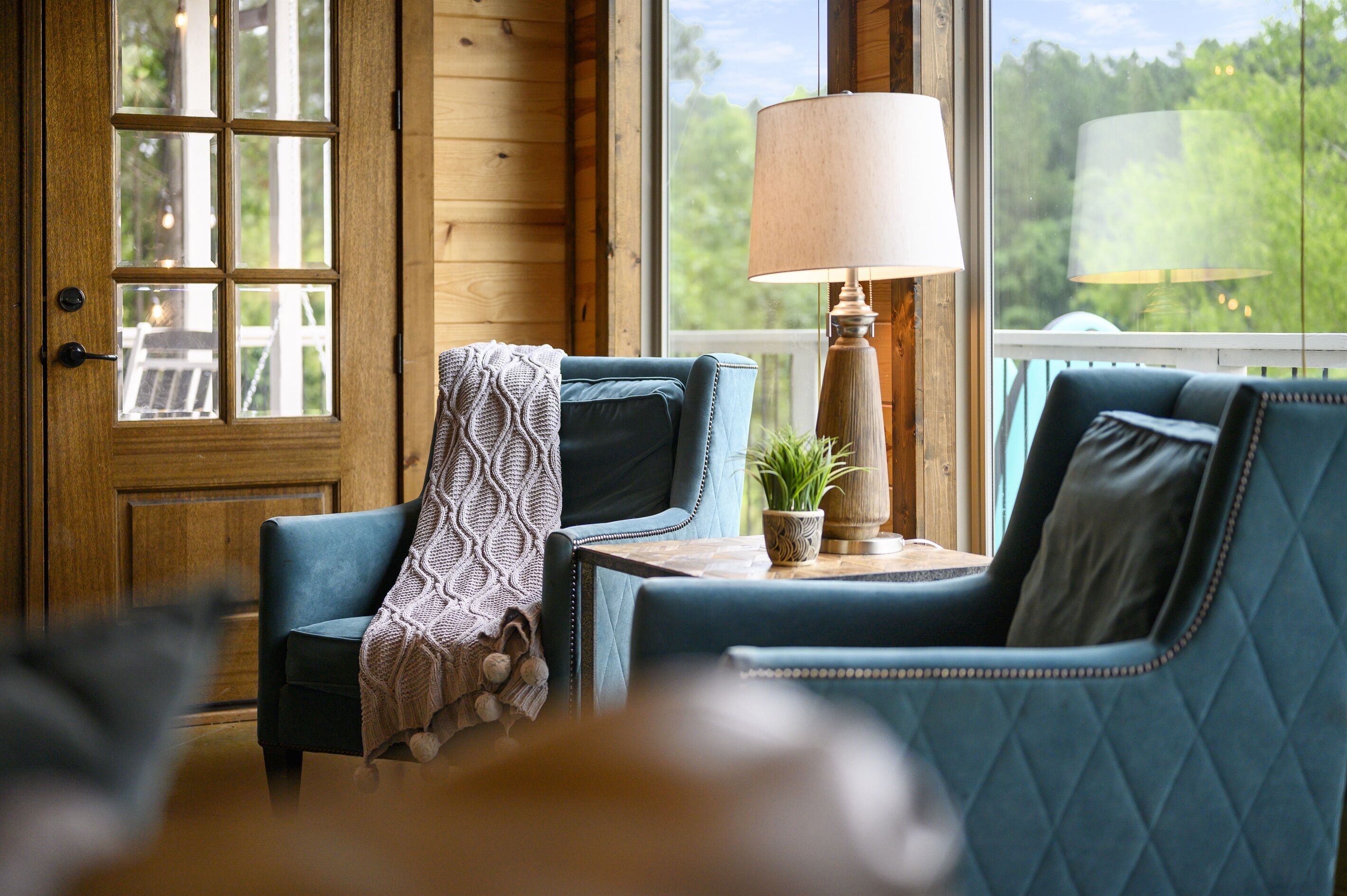 Une pièce chaleureuse avec une porte en bois, un fauteuil bleu, une petite table avec une lampe et une vue sur la verdure à l’extérieur.